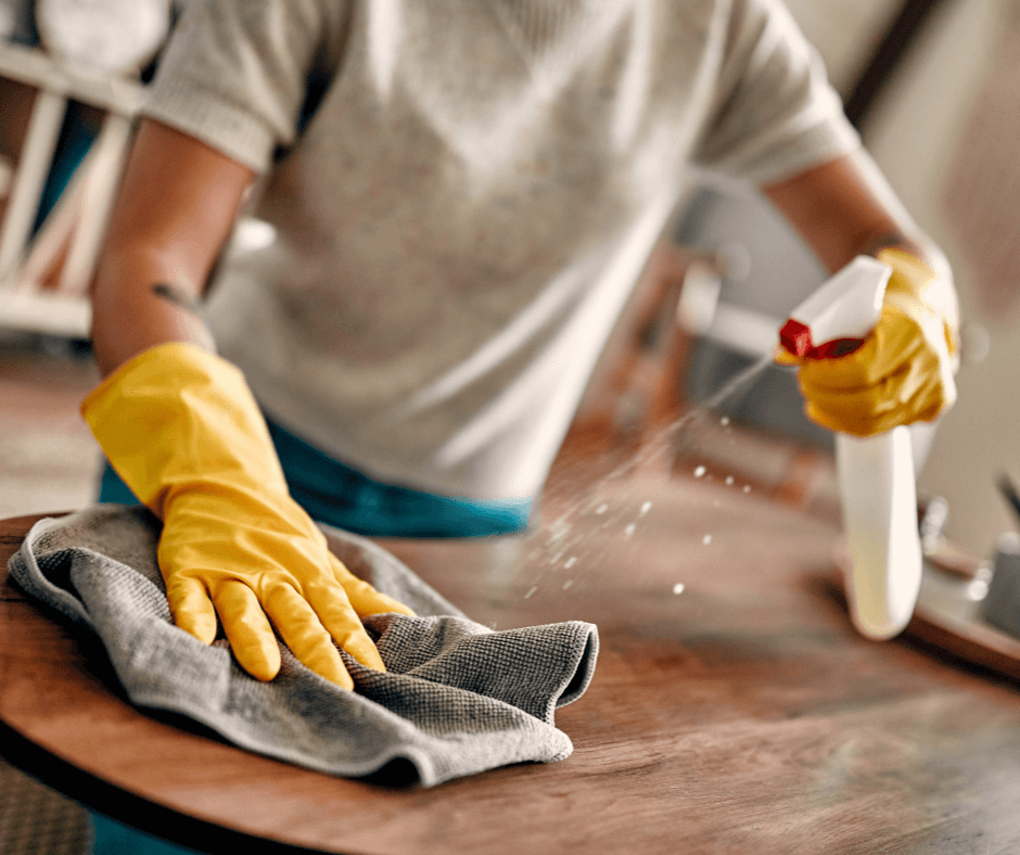 Cleaner wearing yellow gloves spraying disinfectant and wiping wooden table during professional end of tenancy cleaning service in London.